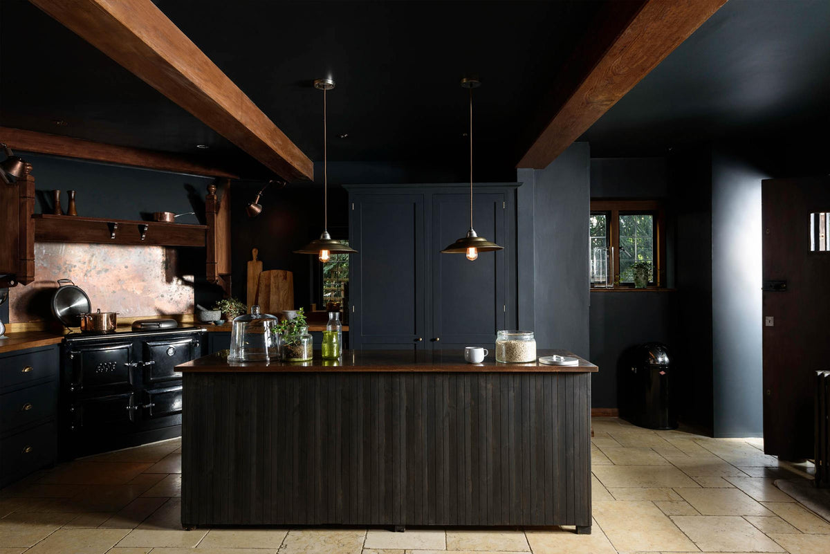 A sophisticated Pantry Blue shaker kitchen with a cooker and sink run, along with a vintage island topped with Iroko wood. Light stone flooring enhances the space, featuring wooden ceiling accents, a large cooker splashback, and two hanging lights above the island.