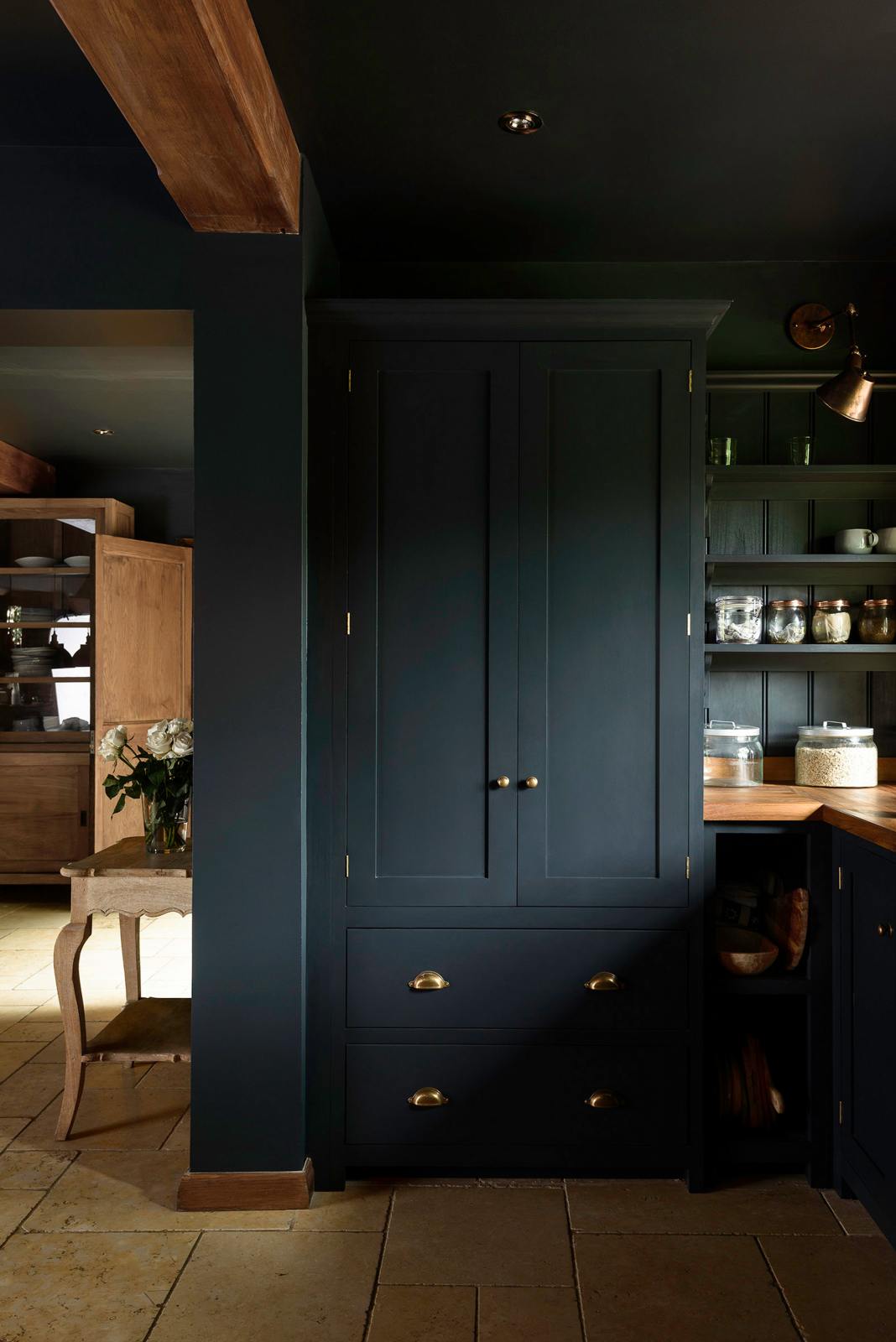 A closer look at the tall Pantry Blue double-door pantry cupboard, featuring open shelving and a cabinet with an Iroko wood worktop. The light stone flooring enhances the space, complemented by wooden ceiling accents.