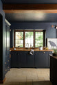 The Pantry Blue shaker kitchen features a sink run that is complemented by a vintage island with an Iroko wood countertop. Light stone flooring enhances the space, which includes wooden ceiling accents. The kitchen overlooks a large window that offers a stunning view of the surrounding trees and foliage.