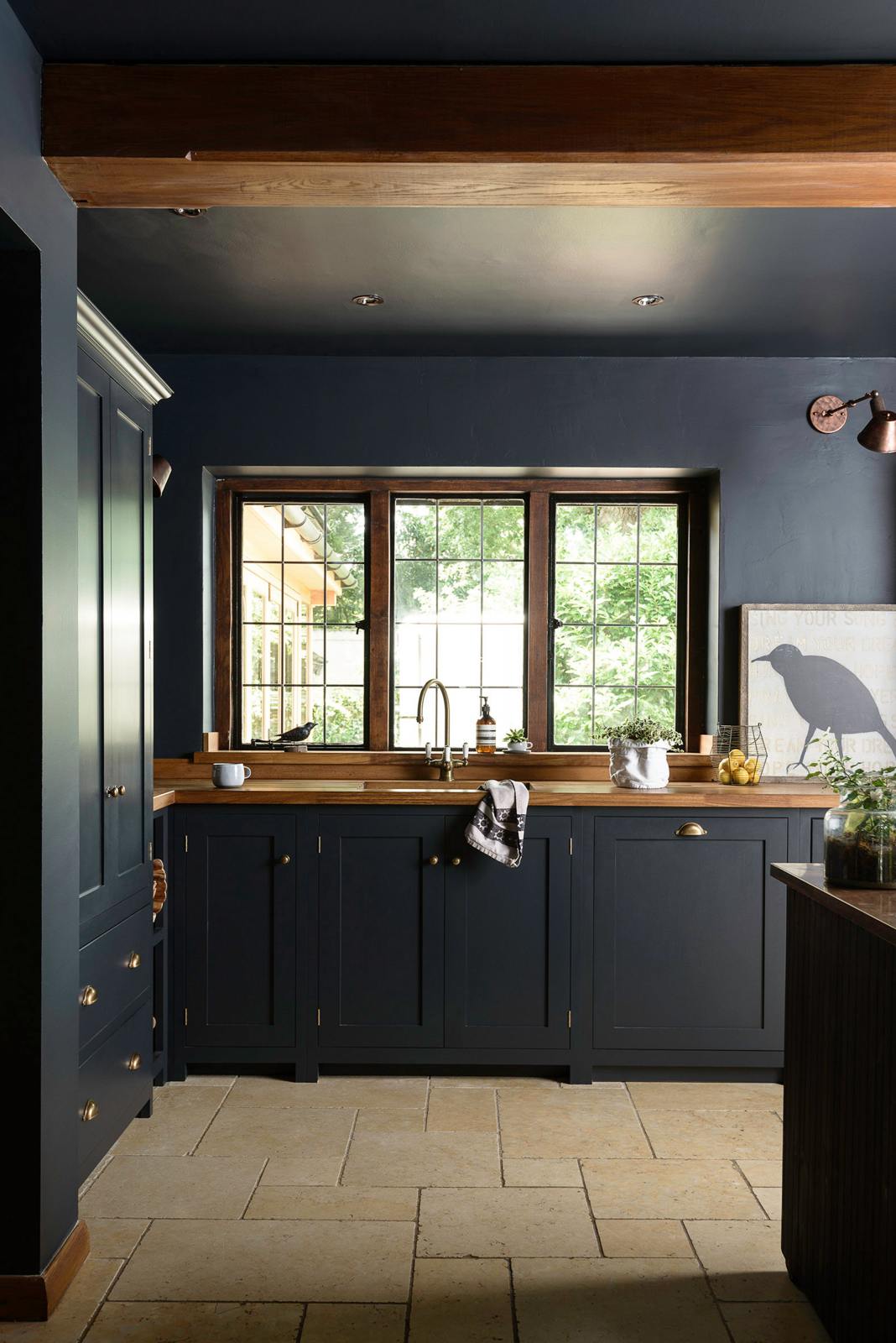 The Pantry Blue shaker kitchen features a sink run that is complemented by a vintage island with an Iroko wood countertop. Light stone flooring enhances the space, which includes wooden ceiling accents. The kitchen overlooks a large window that offers a stunning view of the surrounding trees and foliage.