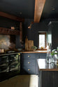 A detailed view of the Pantry Blue shaker kitchen features a cooker run with an Iroko wood worktop. Light stone flooring enhances the space, complemented by wooden ceiling accents, a large cooker splashback, and two hanging lights above the island.