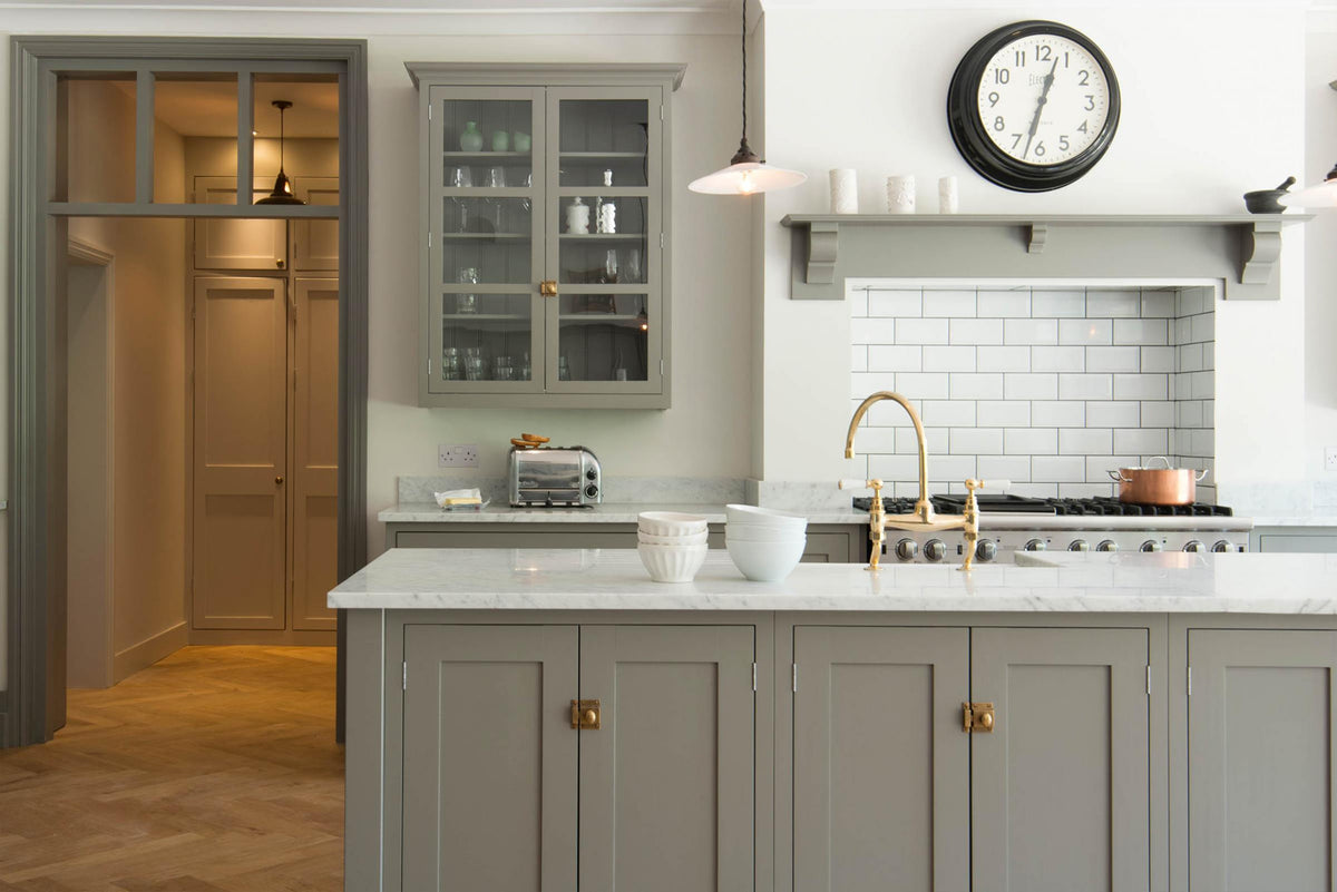 A grand warm-grey Shaker kitchen features a cooker run with a Carrara marble worktop, glazed wall cabinets, an overmantle canopy, and a wide kitchen island with a single sink. Adorned with beloved furniture, simple white platters, glass pendant lights, and pale wood herringbone floors, the space feels sophisticated and cool.