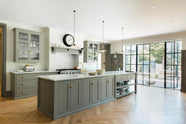 An angled view of a grand warm-grey Shaker kitchen features a cooker run with a Carrara marble worktop, glazed wall cabinets, an overmantle canopy, and a wide kitchen island with a single sink. Adorned with beloved furniture, simple white platters, glass pendant lights, and pale wood herringbone floors, the space feels sophisticated and cool.