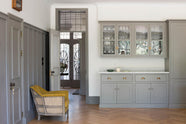 A quiet corner of a grand warm-grey Shaker kitchen features a pantry run with a Carrara marble worktop and glazed wall cabinets. Adorned with beloved furniture, simple white platters, a yellow armchair, and pale wood herringbone floors, the space feels effortlessly sophisticated and cool.