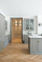 A quiet corner of a grand warm-grey Shaker kitchen features a cooker run with a Carrara marble worktop, glazed wall cabinets, an overmantle canopy, a wide kitchen island with a single sink, and a pantry run. Adorned with beloved furniture, simple white platters, a yellow armchair, and pale wood herringbone floors, the space feels effortlessly sophisticated and cool.
