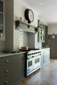 An angled view of a grand warm-grey Shaker kitchen features a cooker run with a Carrara marble worktop, glazed wall cabinets, and an overmantle canopy. Adorned with beloved furniture, simple white platters, a large black-rimmed clock, and pale wood herringbone floors, the space feels sophisticated and cool.