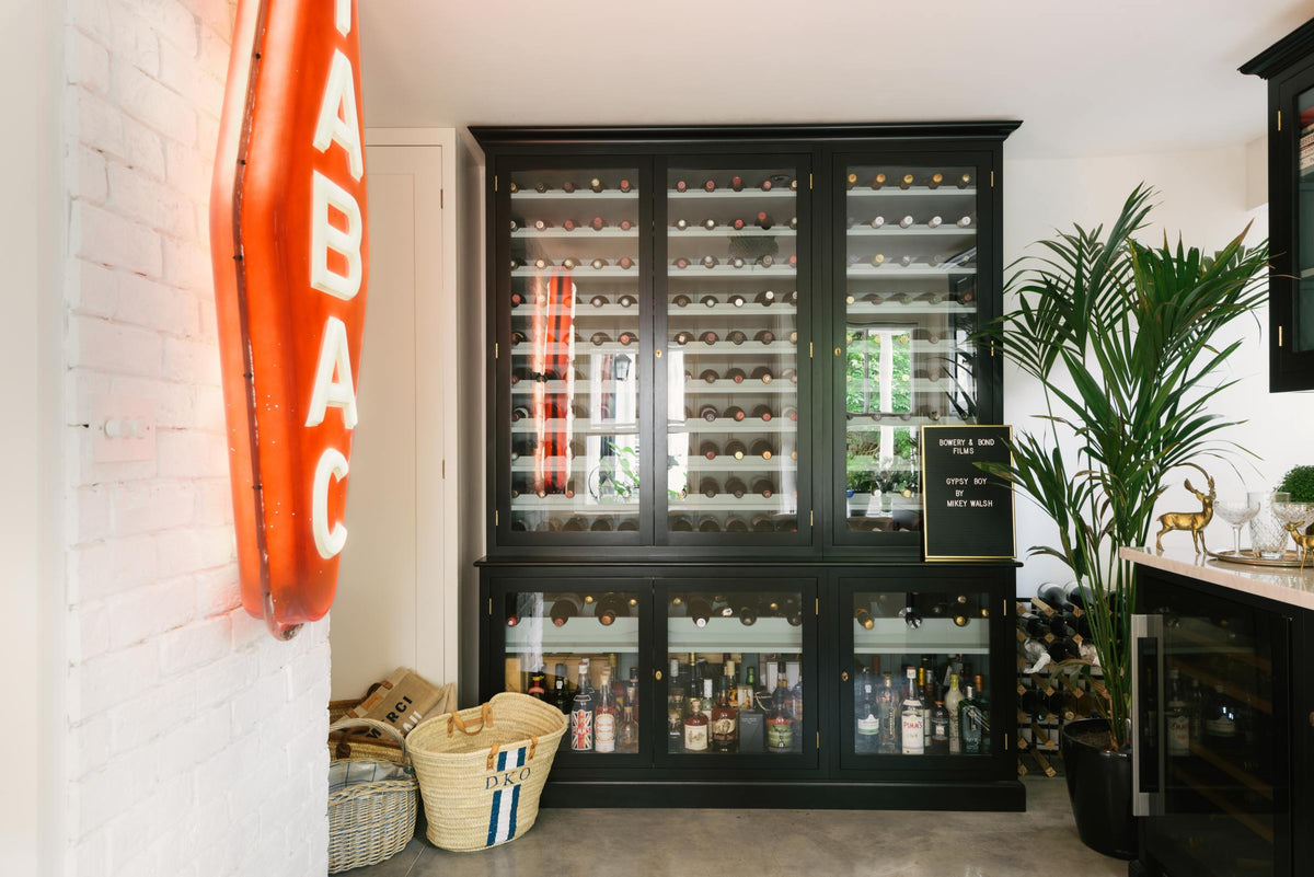A classy cellar bar features a bank of curiosity cupboards, glazed wall cupboards, and a small wine cooler run with a Carrara marble worktop. Adorned with golden embellishments such as deer ornaments, serving trays, and a vintage red Tabac neon lights, it creates a sophisticated and social space.