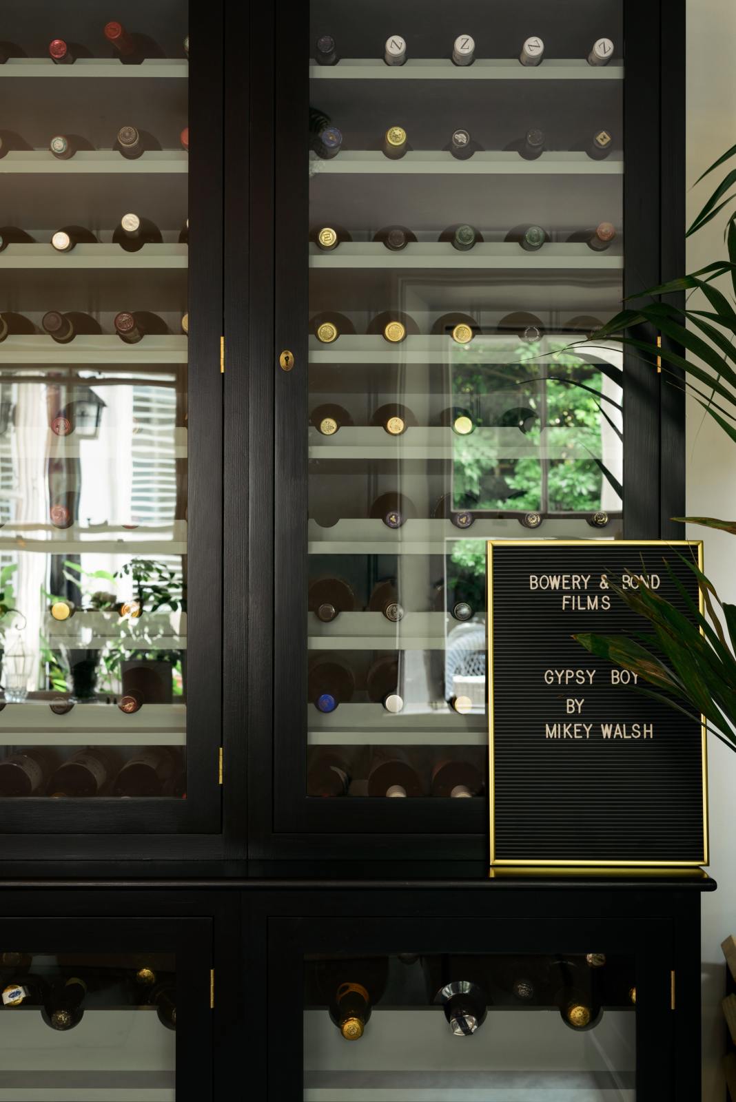 A closer look at a classy cellar bar’s curiosity cupboards reveals a vintage wine collection resting on wine racks. Adorned with a tall house plant and a golden-framed peg letter board, it creates a sophisticated and social space.