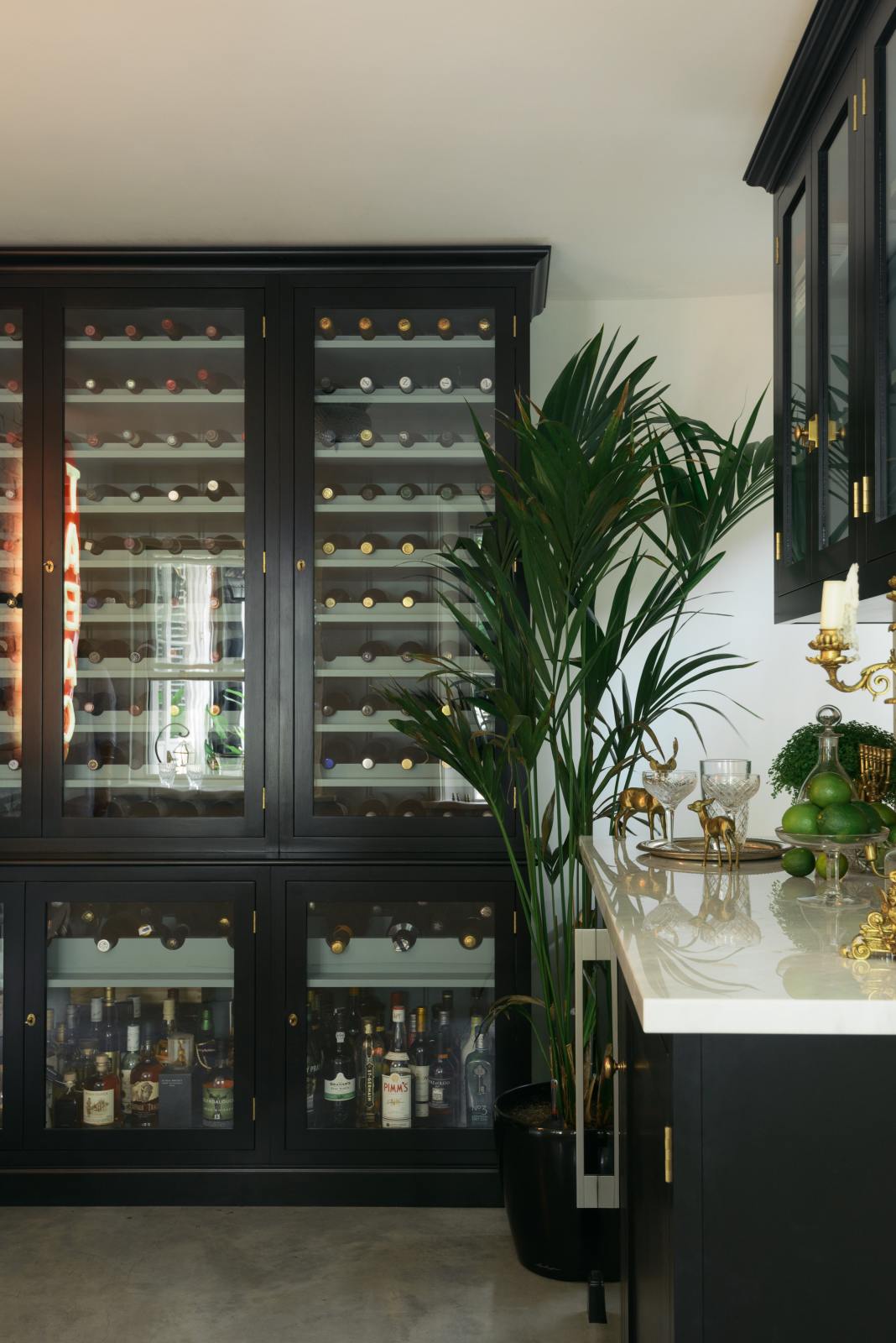 A classy cellar bar features a bank of curiosity cupboards, glazed wall cupboards, and a small wine cooler run with a Carrara marble worktop. Adorned with golden embellishments such as candelabras, a cake stand, deer ornaments, and a golden-framed gentleman's silhouette illustration, it creates a sophisticated and social space.