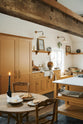 An oblique view of a scullery yellow shaker kitchen with a small sink run with a Carrara marble sink, a full-height cabinet, shaker shelves, an L-shaped shaker bench seat and a closed larder with glazed cupboards. The kitchen features a collection of vintage furniture and beloved items such as mixing bowls and fresh produce. The ceilings are high and retain their original exposed wooden structure.