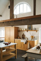 An oblique view of a scullery yellow shaker kitchen with a small sink run with a Carrara marble sink, a full-height cabinet, shaker shelves, an L-shaped shaker bench seat and a kitchen island. The kitchen features a single Belfast sink with aged brass Mayan taps, a collection of vintage furniture and beloved items. The ceilings are high and retain their original exposed wooden structure.