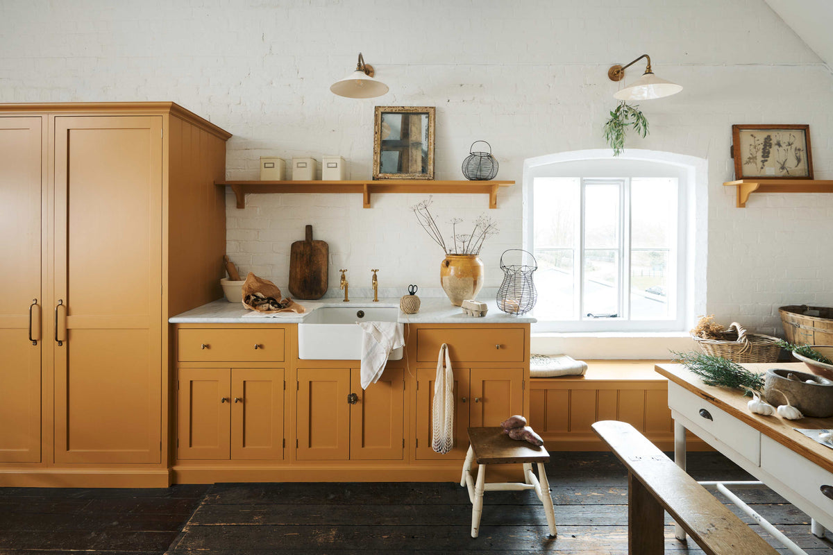 A scullery yellow shaker kitchen with a small sink run with a Carrara marble sink, a full-height cabinet, shaker shelves and a shaker bench seat. The kitchen features a single Belfast sink with aged brass Mayan taps, a collection of vintage furniture and beloved items.