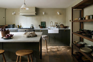 An olive green Shaker kitchen featuring a sink and cooker run, a freestanding prep table with a Carrara marble worktop, and Helen Stools gathered around the breakfast nook. A large wooden shelf displays a thoughtful selection of muted ceramics and mugs. The space embraces a simple theme rooted in raw, natural materials—an ode to Mother Nature—highlighted by a small oil painting of trees placed beside the single Belfast sink with an aged brass Ionian tap.