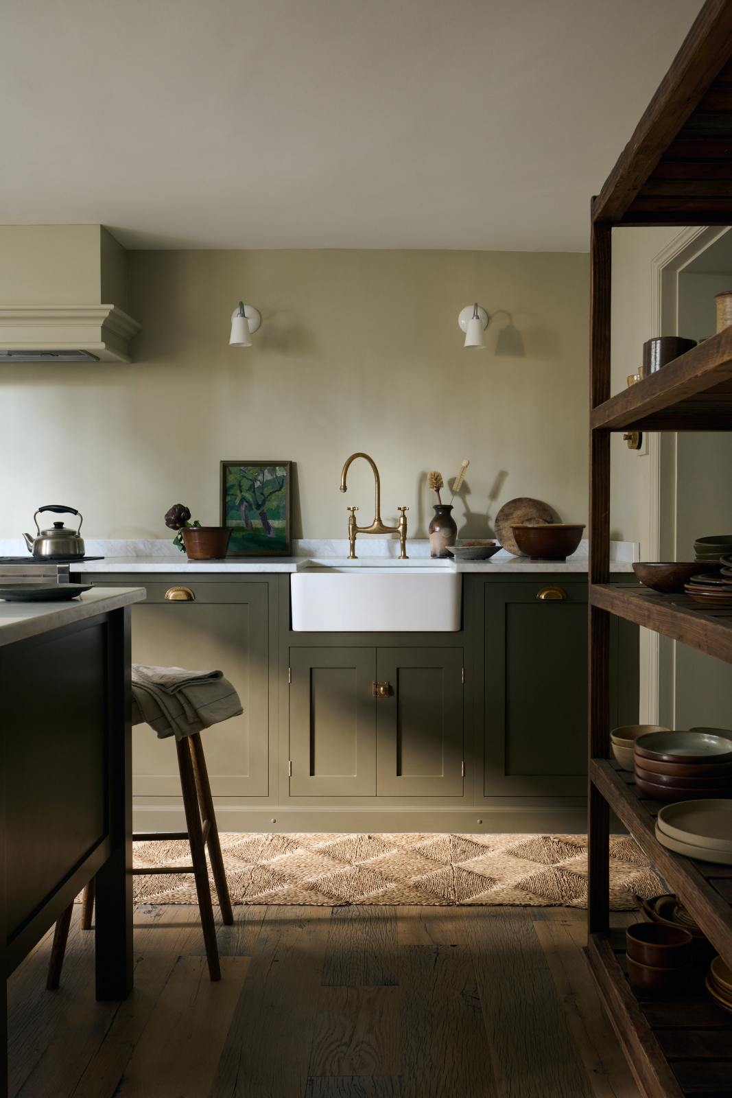 A closer look at an olive green Shaker kitchen featuring a sink and cooker run, a freestanding prep table with a Carrara marble worktop, and Helen Stools gathered around the breakfast nook. A large wooden shelf displays a thoughtful selection of muted ceramics and mugs. The space embraces a simple theme rooted in raw, natural materials—an ode to Mother Nature—highlighted by a small oil painting of trees placed beside the single Belfast sink with an aged brass Ionian tap.