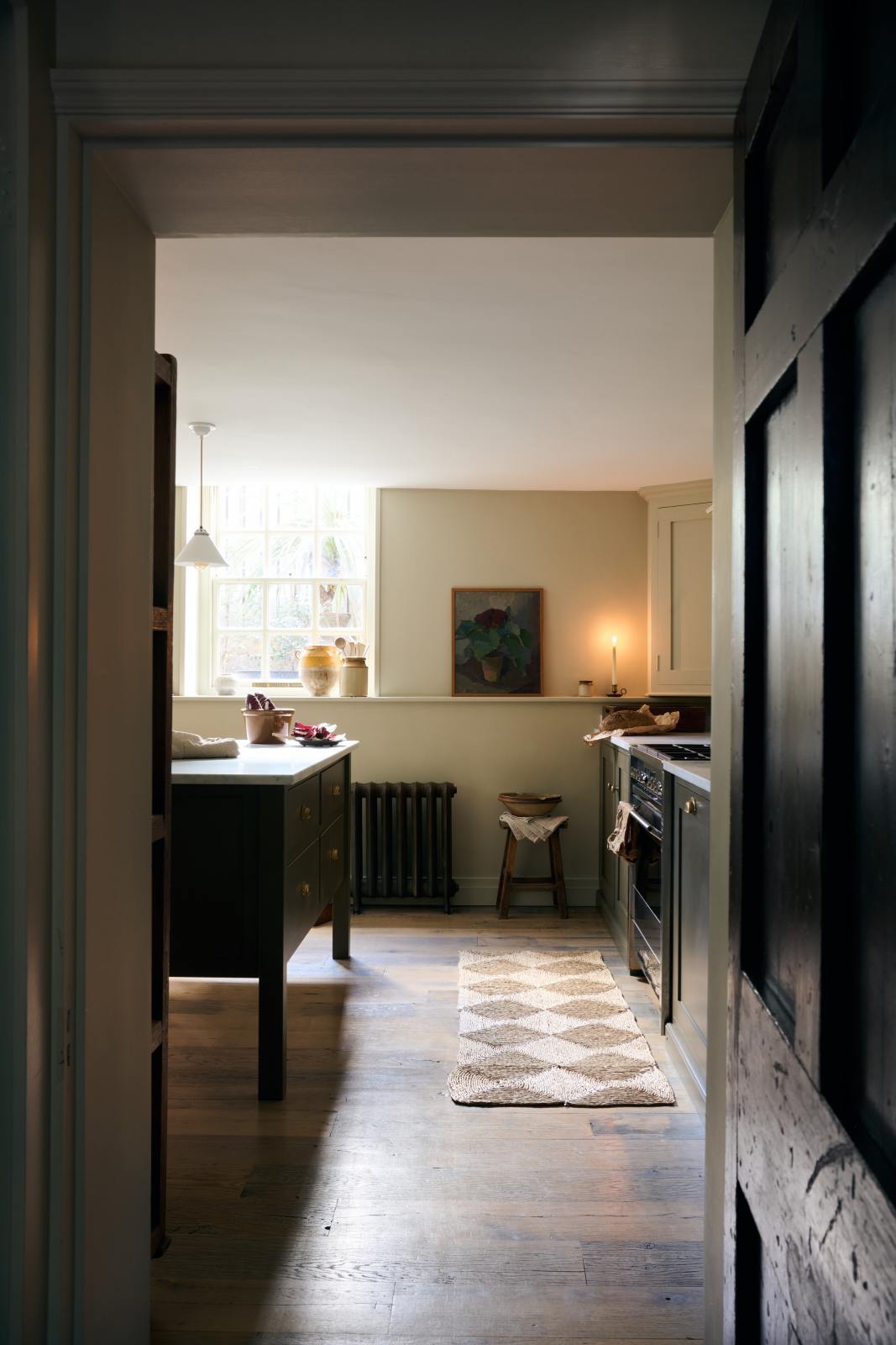 Looking through a dark corridor, a well-lit olive green Shaker kitchen comes into view, featuring a sink and cooker run, along with a freestanding prep table topped with Carrara marble.
