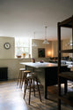 An angled olive green Shaker kitchen featuring a sink and cooker run, a freestanding prep table with a Carrara marble worktop, and Helen Stools gathered around the breakfast nook. A large wooden shelf displays a thoughtful selection of muted ceramics and mugs. The space embraces a simple theme rooted in raw, natural materials—an ode to Mother Nature—highlighted by a small oil painting of trees placed beside the single Belfast sink with an aged brass Ionian tap.