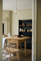 Looking through the open white door, a pale wooden dining table with matching chairs comes into view. Behind it, a freestanding open larder displays beautifully simple cream, green, and reddish-brown ceramic vases and jugs.