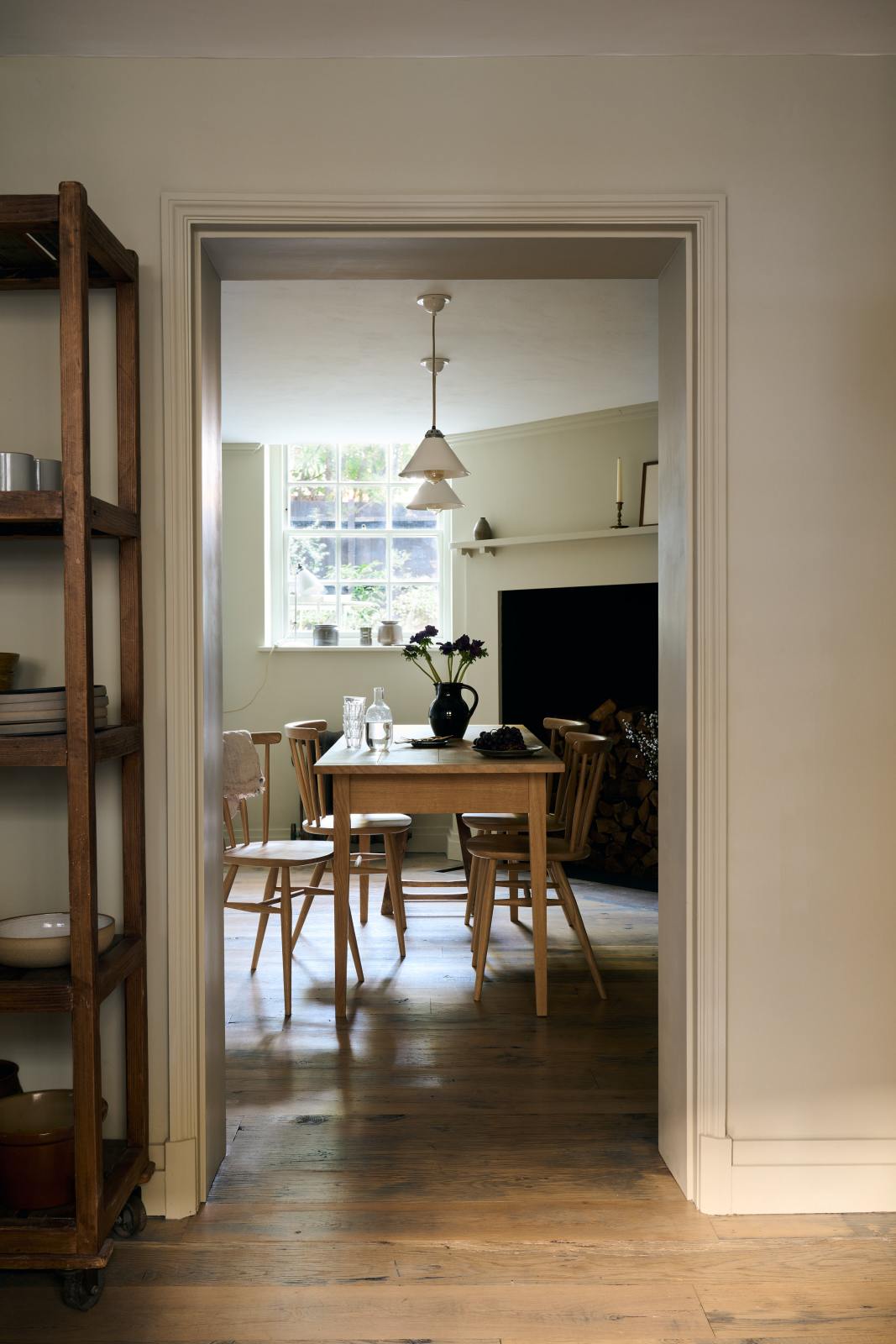 Looking through the archway in the kitchen, beside a large wooden shelf displaying a thoughtful selection of muted ceramics and mugs, the dining room comes into view with a pale wooden dining table and matching chairs. A large open fireplace adds warmth to the space, accompanied by a generous storage cupboard and cabinets. A freestanding open larder showcases beautifully simple cream, green, and reddish-brown ceramic vases and jugs.