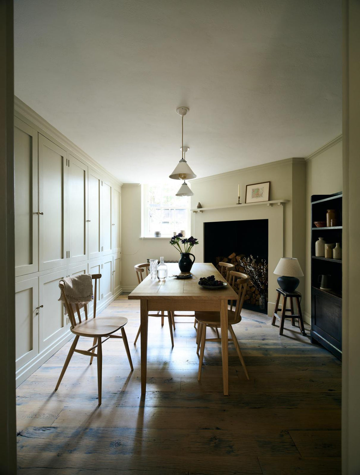 Looking through the archway in the kitchen, beside a large wooden shelf displaying a thoughtful selection of muted ceramics and mugs, the dining room comes into view with a pale wooden dining table and matching chairs.