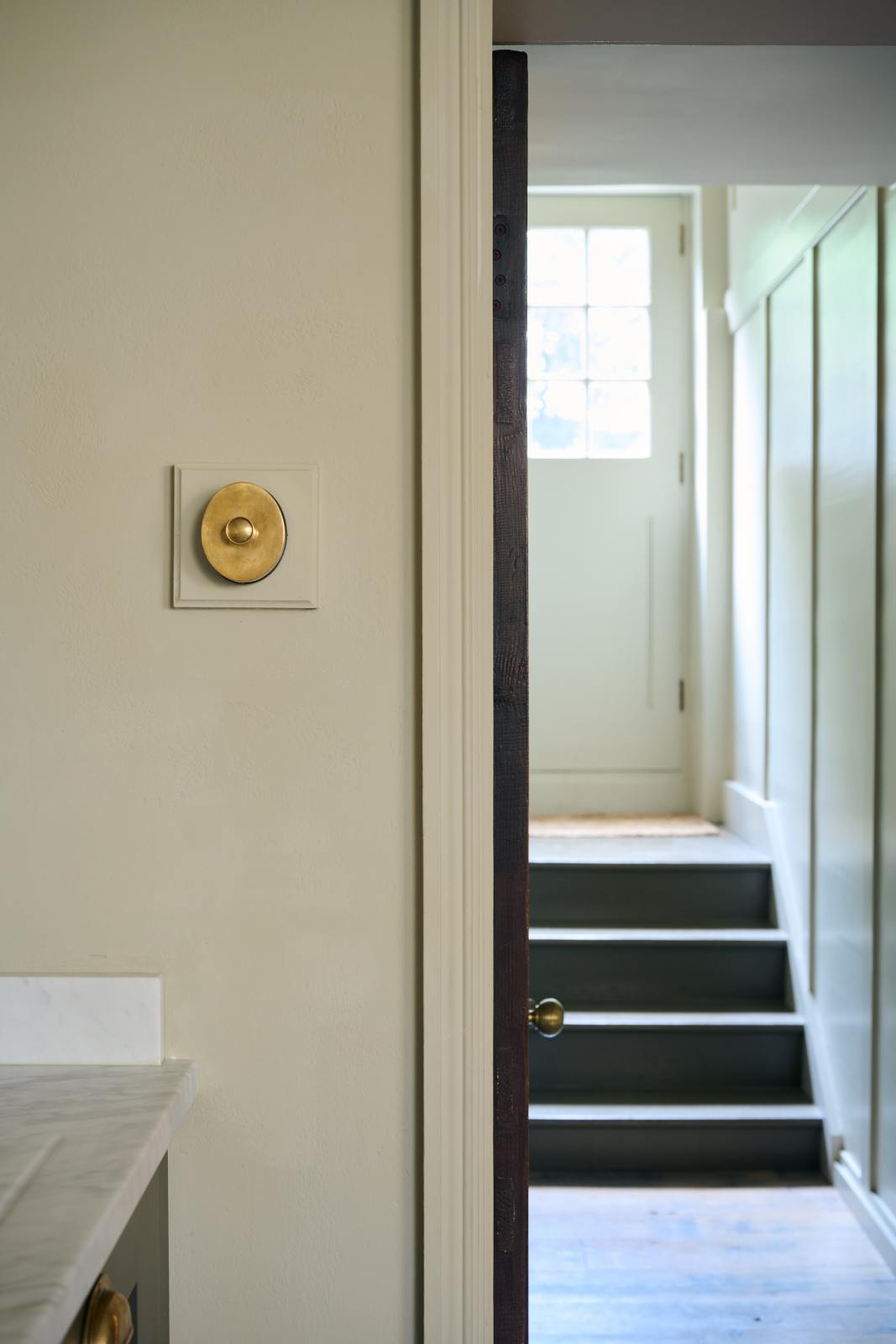 A close-up shot of a deVOL’s petite oval dimmer switch in a traditional and warm heirloom brass finish, fitted in a simple basement kitchen of a Georgian townhouse. There is a beautiful contrast between the glow of the brass dimmer switch and the earthy tones of the greeny-beige painted walls.