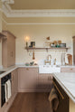Past the large kitchen island of a romantic and peaceful Princelet pink Shaker kitchen is a tactile corner of a sink run, a countertop cupboard, shelf with brass railings, textured marble worktop, and a fridge and freezer cupboard. Beautifully paired with the subtle butter yellow painted walls, decorative cornice and assorted vintage personal items, it creates a warm and inviting space.