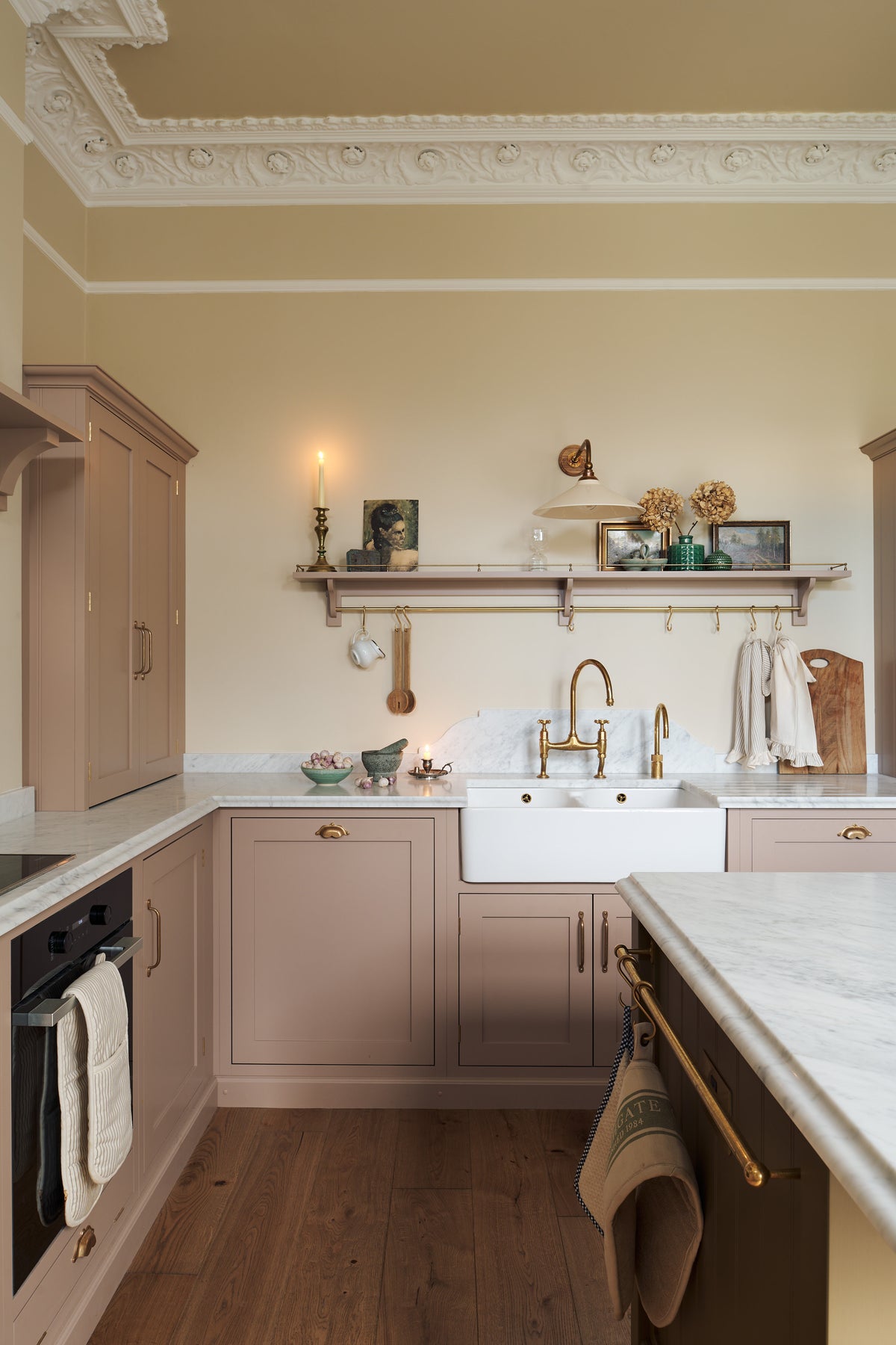 Past the large kitchen island of a romantic and peaceful Princelet pink Shaker kitchen is a tactile corner of a sink run, a countertop cupboard, shelf with brass railings, textured marble worktop, and a fridge and freezer cupboard. Beautifully paired with the subtle butter yellow painted walls, decorative cornice and assorted vintage personal items, it creates a warm and inviting space.