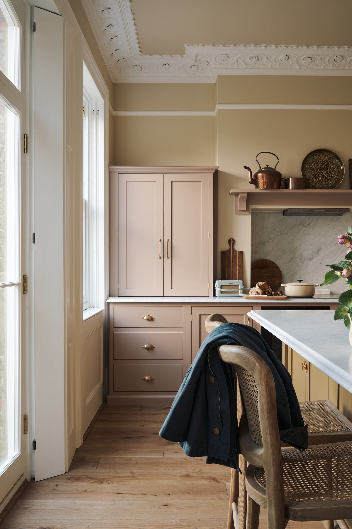 Past the large kitchen island of a romantic and peaceful Princelet pink Shaker kitchen is a quiet corner of a cooker run, a countertop cupboard, a marble worktop, an overmantle above the chimney breast, and a large white window sitting peacefully beside it. Beautifully paired with the subtle butter yellow painted walls, decorative cornice and assorted vintage personal items, it creates a warm and inviting space.