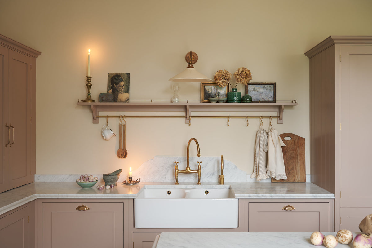 Past the large kitchen island of a romantic and peaceful Princelet pink Shaker kitchen is a tactile corner of a sink run, a countertop cupboard, shelf with brass railings, textured marble worktop, and a fridge and freezer cupboard. Beautifully paired with the subtle butter yellow painted walls, decorative cornice and assorted vintage personal items, it creates a warm and inviting space.
