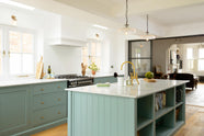 A bright and elegant Trinity blue Shaker kitchen features a long cooker run with a canopy, a kitchen island with a single sink, an aged brass Ionian tap, and a Carrara marble worktop. Beautifully decorated with artisan ceramics, books, wooden chopping boards, and a watermelon, it creates a humble family space.