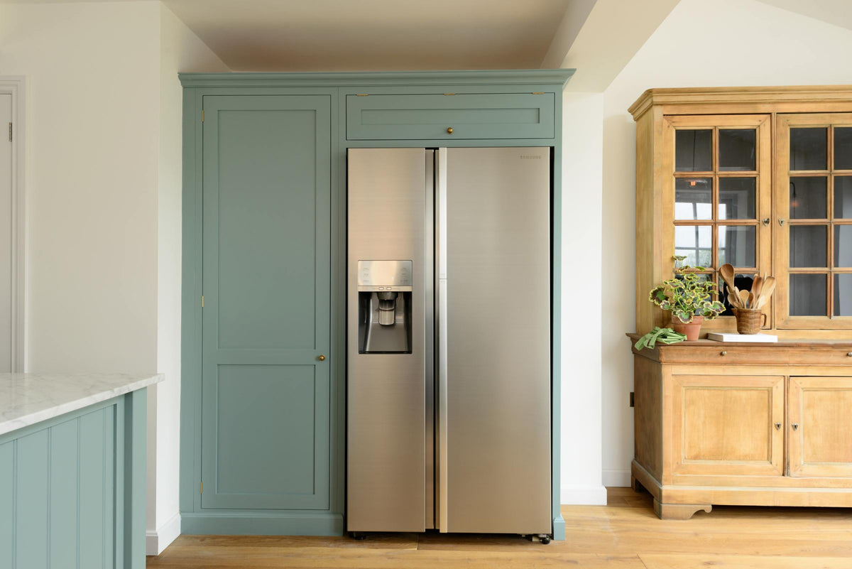 A bright and elegant Trinity blue Shaker kitchen features a full-height cupboard, a silver refrigerator, and a vintage dresser. Beautifully decorated with artisan ceramics, books, wooden utensils, and a vibrant house plant, it creates a humble family space.