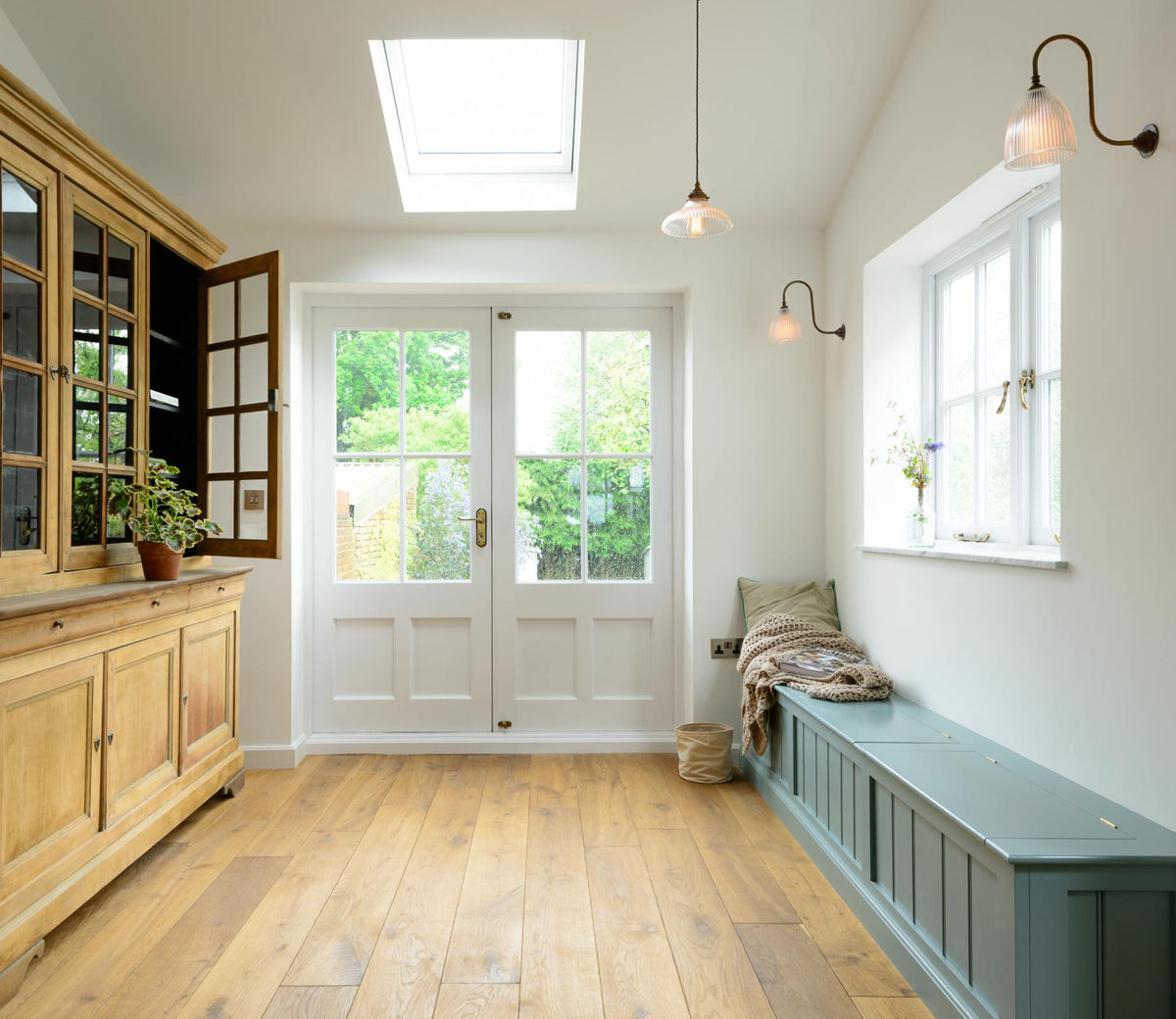 A bright and elegant hallway to the garden features a vintage dresser and a long Shaker bench seat with storage compartments. Beautifully decorated with a vintage dresser, a glass-frilled pendant light, a swan-neck glass-frilled wall light, a pillow, a crocheted blanket, and a vibrant house plant, it creates a humble family space.