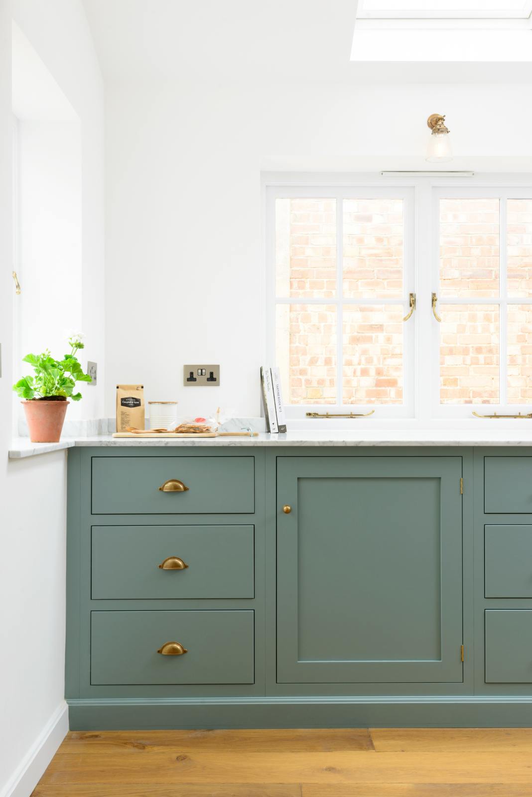 A corner of a bright and elegant Trinity blue Shaker kitchen features a long cooker run with a Carrara marble worktop. Beautifully decorated with artisan coffee beans, books, wooden chopping boards, crackers, and a luscious green house plant, it creates a humble family space.