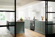 Past a black-framed internal glass sliding door is a bright and elegant Trinity blue Shaker kitchen featuring a long cooker run with a canopy, a kitchen island with a single sink, an aged brass Ionian tap, and a Carrara marble worktop. Beautifully decorated with artisan ceramics, books, wooden chopping boards, and a watermelon, it creates a humble family space.