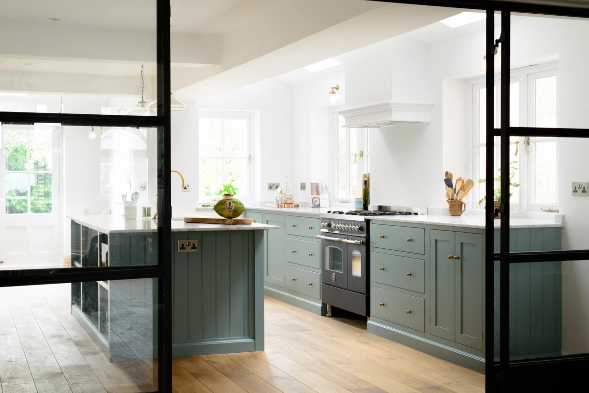 Past a black-framed internal glass sliding door is a bright and elegant Trinity blue Shaker kitchen featuring a long cooker run with a canopy, a kitchen island with a single sink, an aged brass Ionian tap, and a Carrara marble worktop. Beautifully decorated with artisan ceramics, books, wooden chopping boards, and a watermelon, it creates a humble family space.