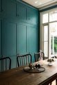 An angled view reveals a grand, seafoam green Shaker kitchen featuring a bank of full-height pantry cupboards with a Carrara marble worktop, and a charming French door leading to the conservatoire. Adorned with beautiful vintage furniture, a glass bowl of green figs, antique silverware, and a decanter, the kitchen reflects the refined tastes of its socialite and collector owners.
