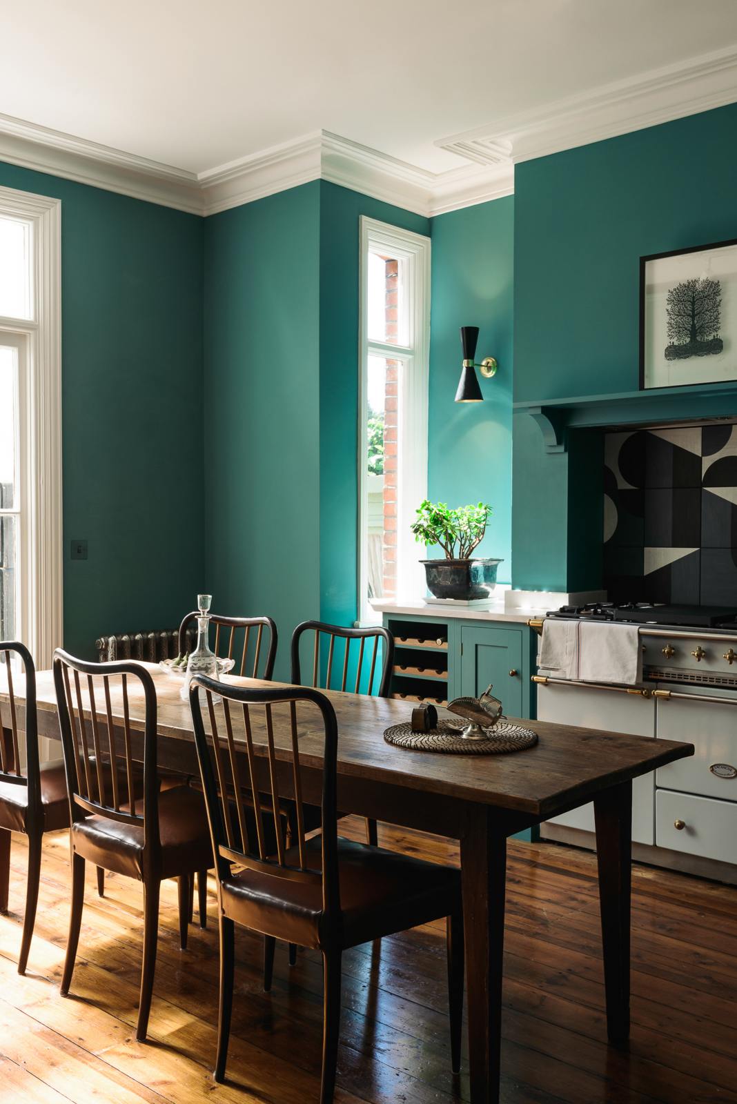 An angled view reveals a grand, seafoam green Shaker kitchen featuring an L-shaped cooker and sink run with a Carrara marble worktop, an overmantle canopy, and abstract splashback tiles. Adorned with beautiful vintage furniture, a luscious green house plant, a detailed block print, antique silverware, and a decanter, the kitchen reflects the refined tastes of its socialite and collector owners.