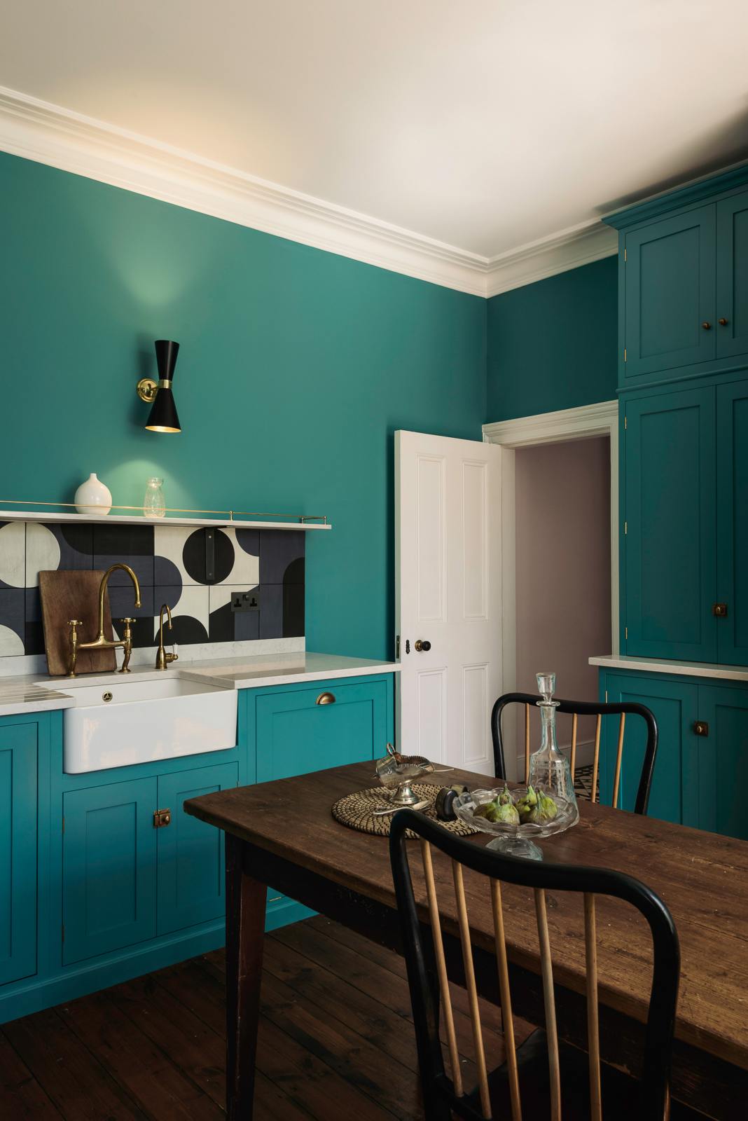 An angled view reveals a grand, seafoam green Shaker kitchen featuring an L-shaped cooker and sink run with a single Belfast sink, an aged brass Ionian tap, and a free-standing closed larder with Carrara marble worktop. Adorned with beautiful vintage furniture, a glass bowl of green figs, antique silverware, and a decanter, the kitchen reflects the refined tastes of its socialite and collector owners.
