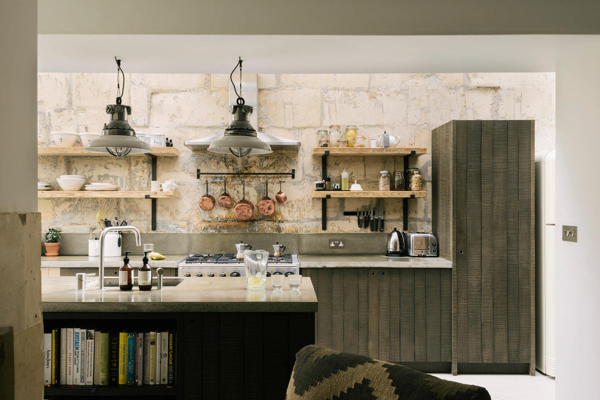 A fresh and calming Sebastian Cox kitchen features a long cooker run with a cement worktop and splashback, a kitchen island with a sink, a full-height larder cupboard, and a stainless steel canopy. Adorned with a bespoke moss green colour tint, copper pans, artisan ceramics, dried ingredients in glass jars, and books, this kitchen creates a practical earth-toned space that complements the original limestone walls.