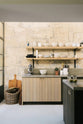 A quiet corner of a fresh and calming Sebastian Cox kitchen features a long cooker run with a cement worktop and splashback, and a kitchen island. Adorned with a bespoke moss green colour tint, large chartreuse board, artisan ceramics, a wicker basket, and a stainless steel mixer, this kitchen creates a practical earth-toned space that complements the original limestone walls.