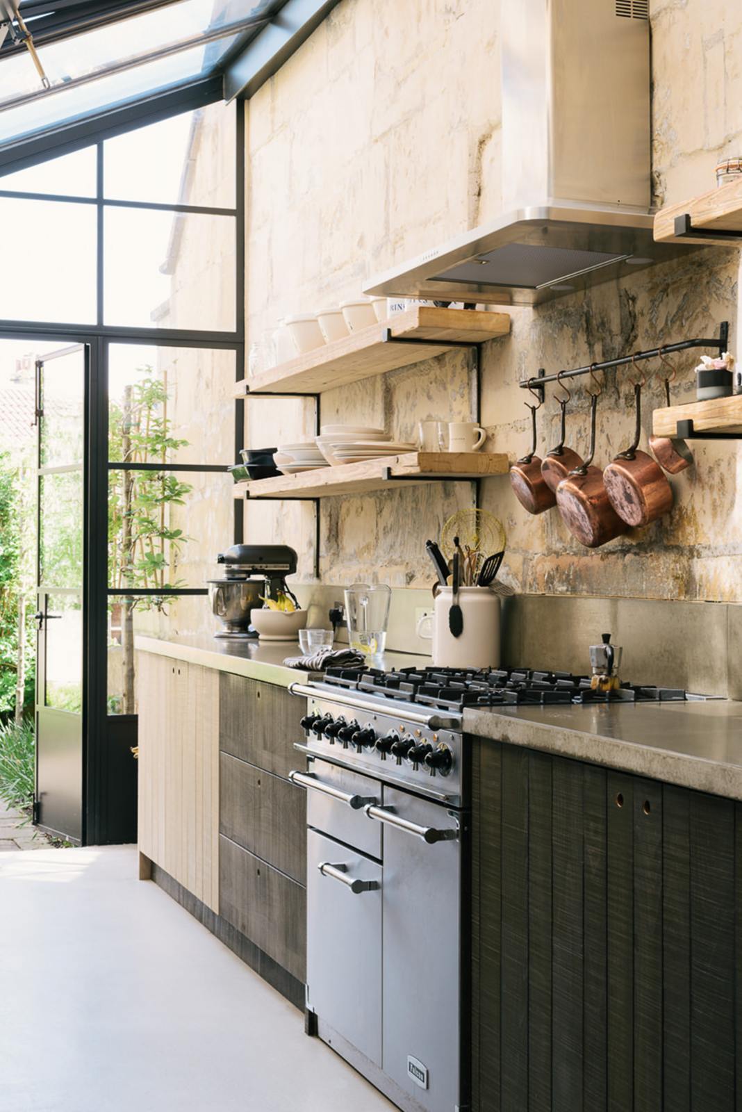 An oblique view of a fresh and calming Sebastian Cox kitchen features a long cooker run with a cement worktop, splashback, and a stainless steel canopy. Adorned with a bespoke moss green colour tint, copper pans, artisan ceramics, and an up-and-over glass design, this kitchen creates a practical earth-toned space that complements the original limestone walls.