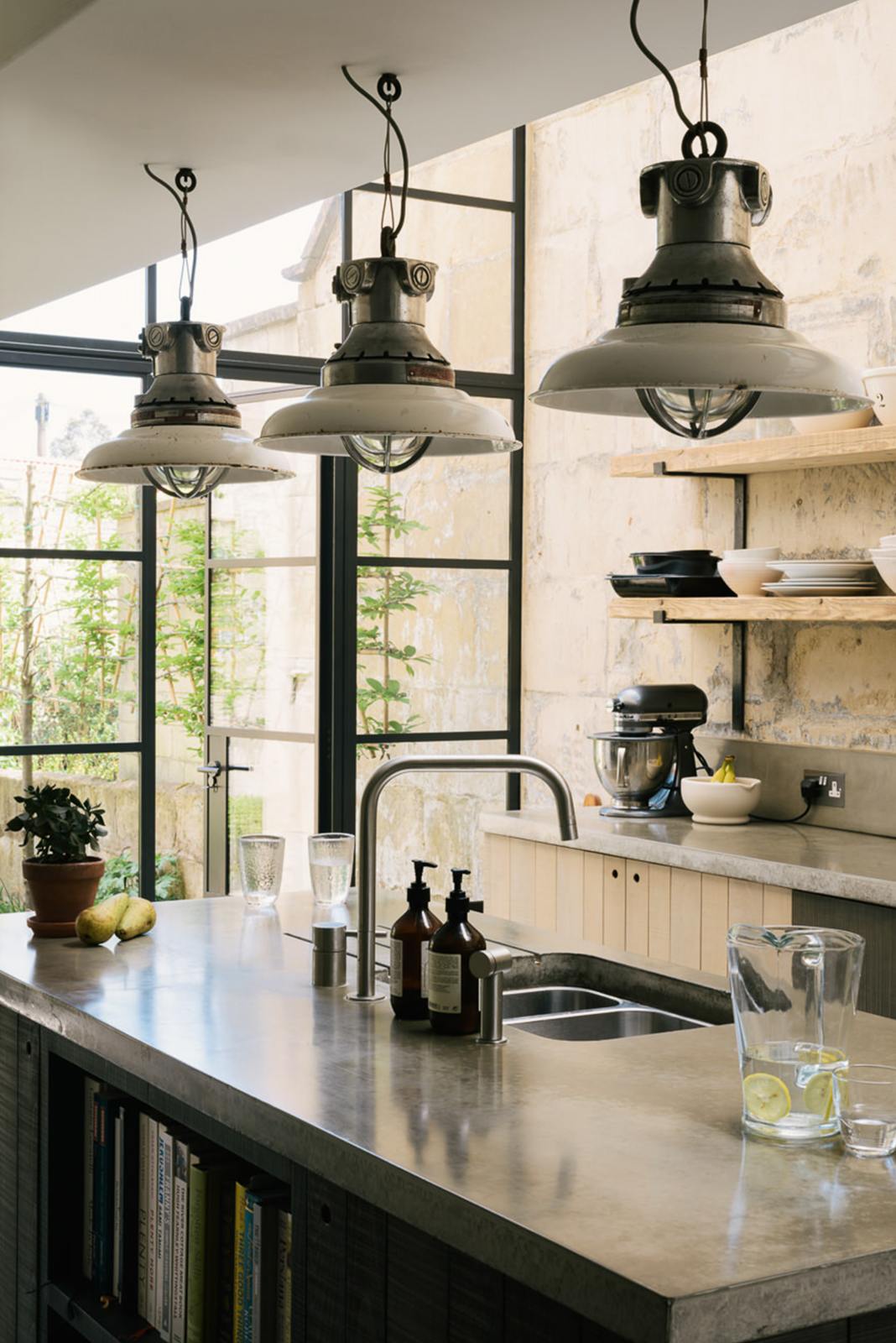 A close-up and oblique view of a fresh and calming Sebastian Cox kitchen features a long cooker run with a cement worktop and splashback, and a kitchen island with a sink. Adorned with a bespoke moss green colour tint, industrial pendant lights, artisan ceramics, pears, bananas, and a jug of fresh lemon water, this kitchen creates a practical earth-toned space that complements the original limestone walls.