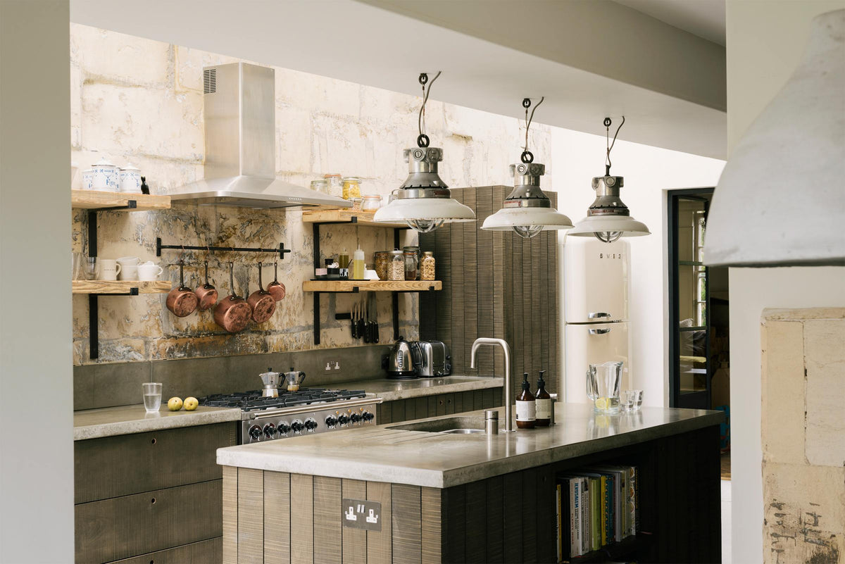 An oblique view of a fresh and calming Sebastian Cox kitchen features a long cooker run with a cement worktop and splashback, a kitchen island with a sink, a full-height larder cupboard, and a stainless steel canopy. Adorned with a bespoke moss green colour tint, industrial pendant lights, copper pans, artisan ceramics, dried ingredients in glass jars, and books, this kitchen creates a practical earth-toned space that complements the original limestone walls.
