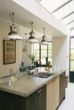 A close-up and oblique view of a fresh and calming Sebastian Cox kitchen features a kitchen island with a stainless steel sink and a cement worktop. Adorned with a bespoke moss green colour tint, industrial pendant lights, pears, and a jug of fresh lemon water, this kitchen creates a practical earth-toned space that complements the original limestone walls.