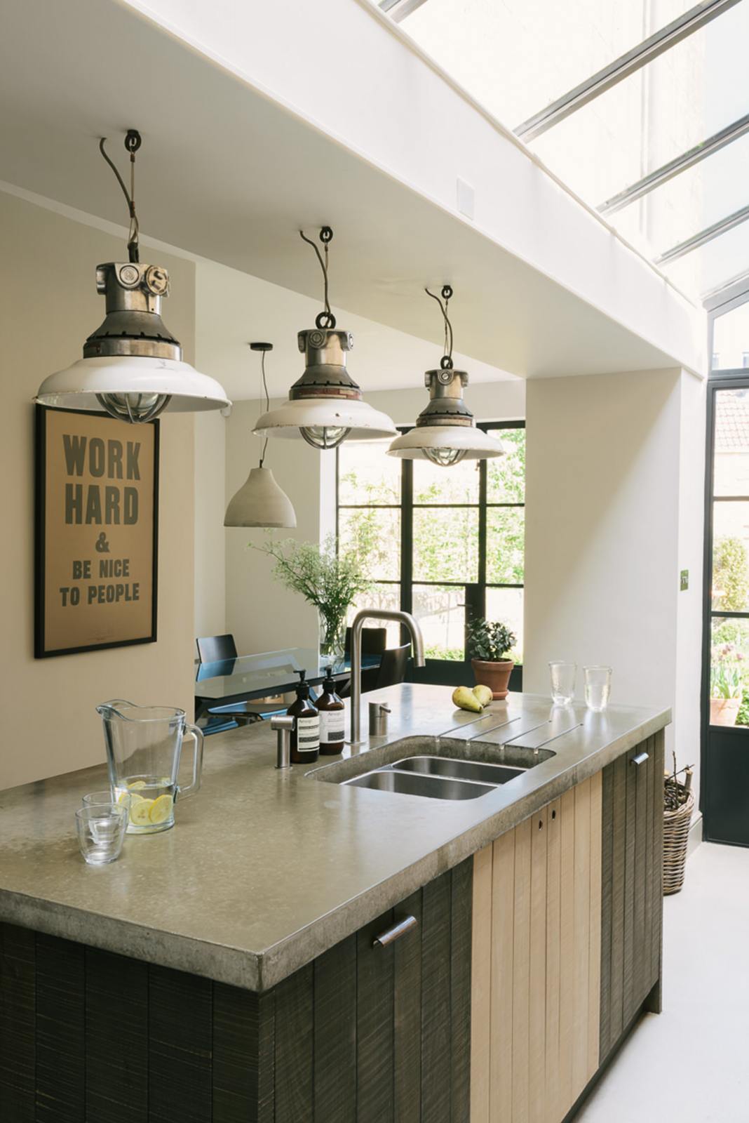 A close-up and oblique view of a fresh and calming Sebastian Cox kitchen features a kitchen island with a stainless steel sink and a cement worktop. Adorned with a bespoke moss green colour tint, industrial pendant lights, pears, and a jug of fresh lemon water, this kitchen creates a practical earth-toned space that complements the original limestone walls.