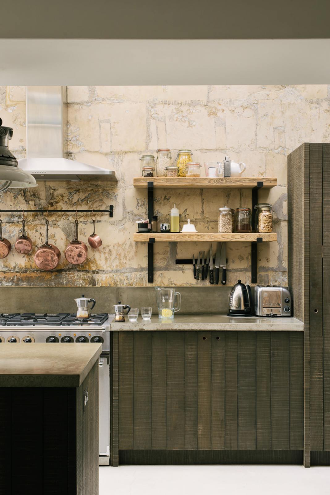 A close-up of a fresh and calming Sebastian Cox kitchen features a long cooker run with a cement worktop and splashback, a kitchen island with a sink, a full-height larder cupboard, and a stainless steel canopy. Adorned with a bespoke moss green colour tint, copper pans, artisan ceramics, dried ingredients in glass jars, and industrial pendant lights, this kitchen creates a practical earth-toned space that complements the original limestone walls.