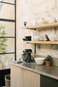 A quiet corner of a fresh and calming Sebastian Cox kitchen features a long cooker run with a cement worktop, splashback, and shelves. Adorned with a bespoke moss green colour tint, artisan ceramics, a wicker basket, a book, and a silver mixer machine, this kitchen creates a practical earth-toned space that complements the original limestone walls.
