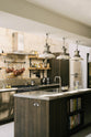 An oblique view of a fresh and calming Sebastian Cox kitchen features a long cooker run with a cement worktop and splashback, a kitchen island with a sink, a full-height larder cupboard, and a stainless steel canopy. Adorned with a bespoke moss green colour tint, copper pans, industrial pendant lights, dried ingredients in glass jars, and books, this kitchen creates a practical earth-toned space that complements the original limestone walls.