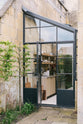 A quaint up-and-over glass design sits between a historic Georgian house, revealing a fresh and calming Sebastian Cox kitchen adorned with a bespoke moss green colour tint, copper pans, and artisan ceramics. This space creates a practical earth-toned environment that complements the original limestone walls.