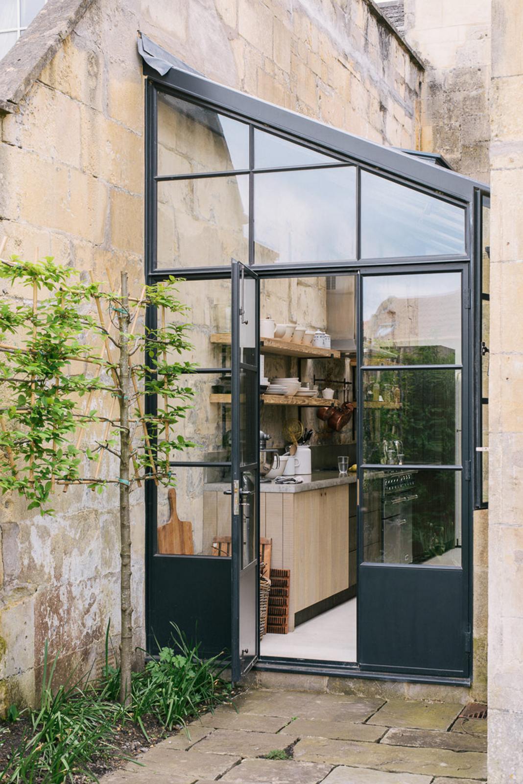 A quaint up-and-over glass design sits between a historic Georgian house, revealing a fresh and calming Sebastian Cox kitchen adorned with a bespoke moss green colour tint, copper pans, and artisan ceramics. This space creates a practical earth-toned environment that complements the original limestone walls.