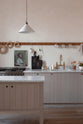 A close-up of a warm and natural Sebastian Cox kitchen features a sink and cooker run with a Carrara marble worktop, a single Belfast sink, an aged brass Ionian tap, a long oak peg rail, and a movable prep table. Beautifully decorated with artisan ceramics, a creamware rise-and-fall light, a plug-in porcelain beaker light, and blackened chopping boards, this kitchen creates a calm and delicate atmosphere that celebrates the use of natural materials.